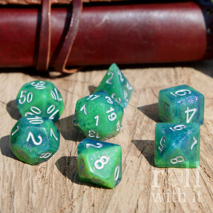 Green blue mixed glitter polyhedral dice set on a wooden surface with a leather-bound book in the background.