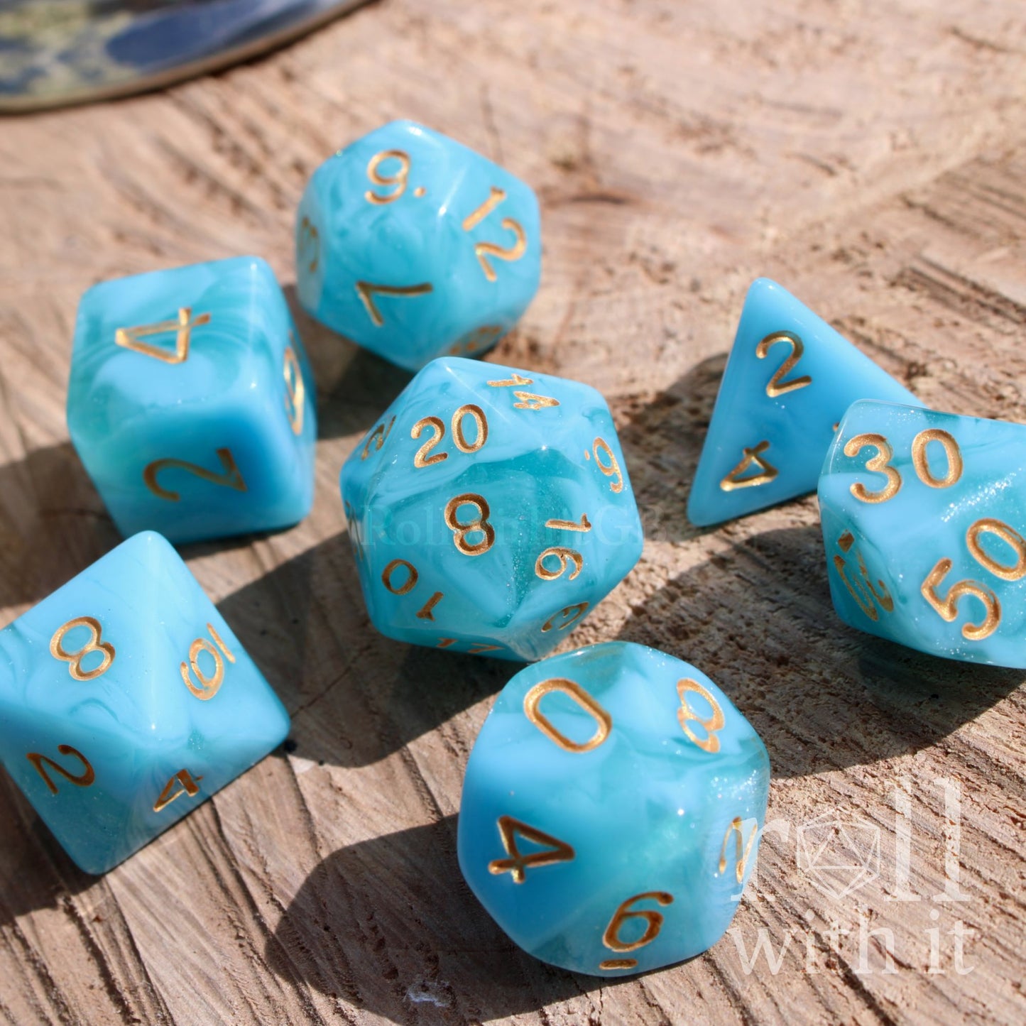 Set of 7 aqua blue polyhedral roleplaying tabletop dice with gold numbers on a wooden surface.