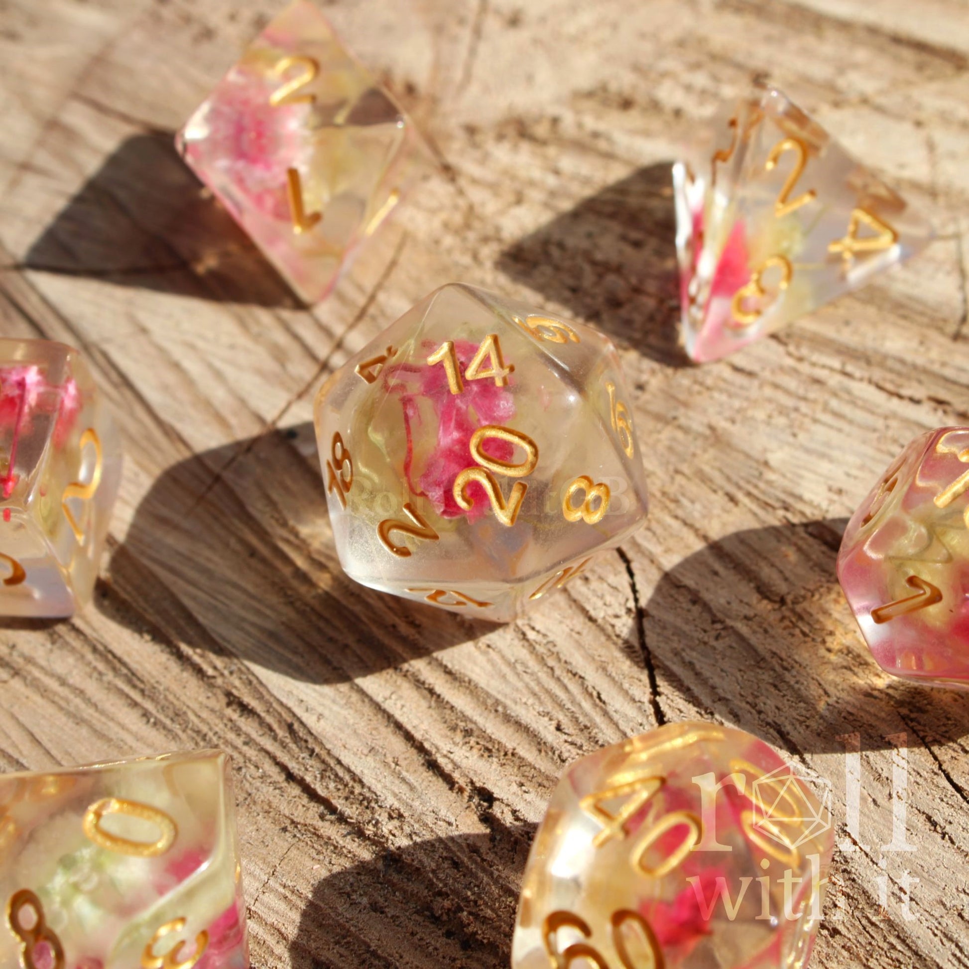 Set of 7 transparent polyhedral roleplaying tabletop dice with pink yellow flowers inside with gold numbers on a wooden surface