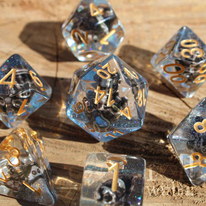 Close up of 7 clear and blue glittery pirate sea themed dungeons and dragons dice with gold numbers on a wooden surface.