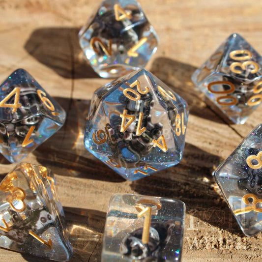 Close up of 7 clear and blue glittery pirate sea themed dungeons and dragons dice with gold numbers on a wooden surface.