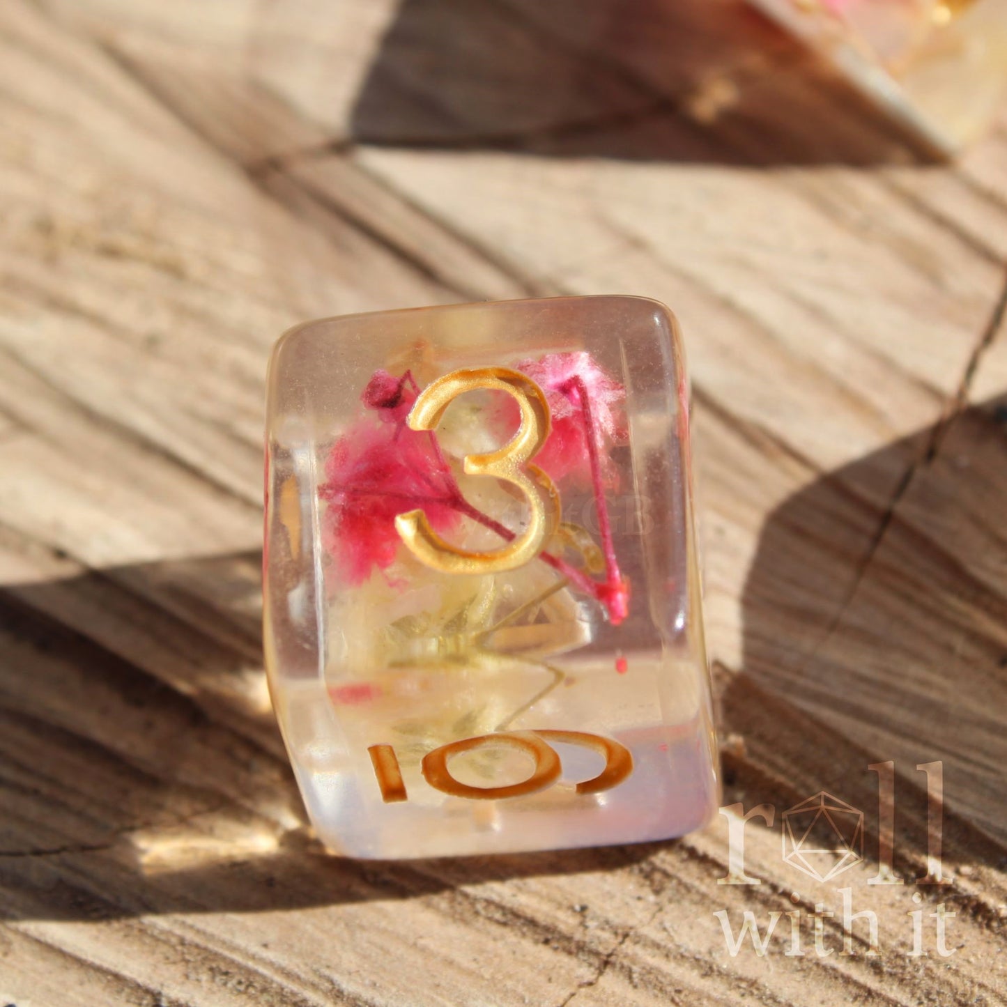 Close up of a single polyhedral TTRPG dice with golden numbers and pink and yellow flowers inside a clear d6 die
