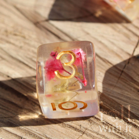 Close up of a single polyhedral TTRPG dice with golden numbers and pink and yellow flowers inside a clear d6 die