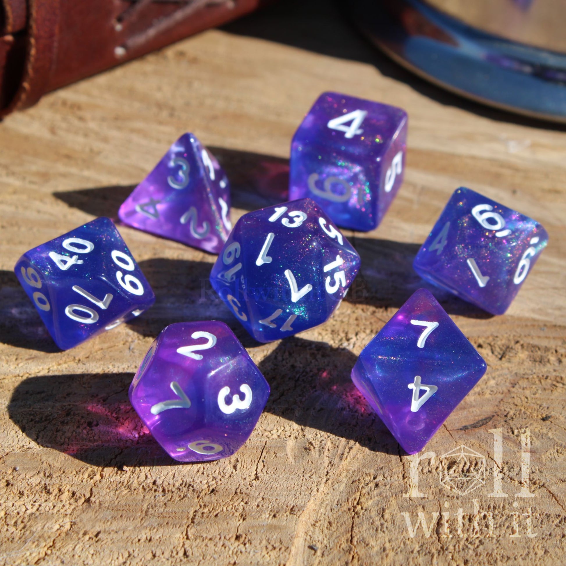 Set of 7 purple and blue galaxy aesthetic polyhedral dice on a wooden surface with a brand logo in the corner.