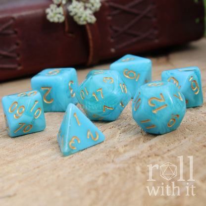 Cute sailor aesthetic set of 7 blue turquoise polyhedral dice set with gold numbers on a wooden surface, featuring the 'roll with it' logo.