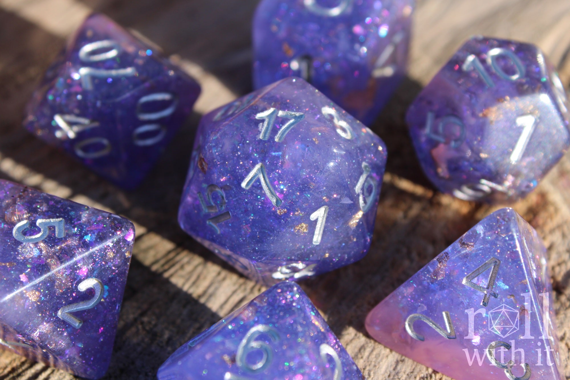Set of purple and pink glittery and gold leaf polyhedral roleplaying dice with silver lettering on a wooden surface