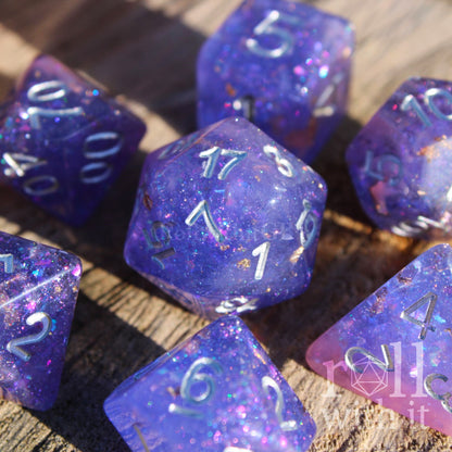 Set of purple and pink glittery and gold leaf polyhedral roleplaying dice with silver lettering on a wooden surface