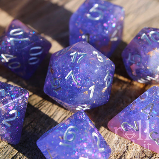 Set of purple and pink glittery and gold leaf polyhedral roleplaying dice with silver lettering on a wooden surface