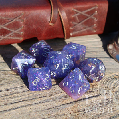 Set of purple pink mixed glitter gold leaf polyhedral dice with silver numbers on a wooden surface, next to a leather-bound book.