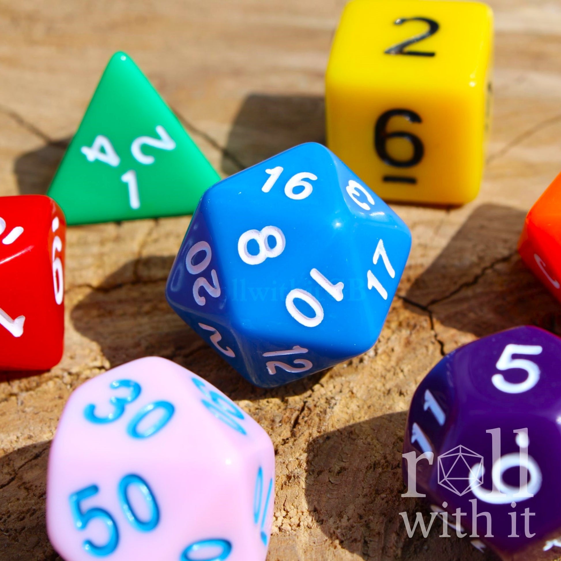 Colorful rainbow blue green yellow orange red pink purple dice with numbers on a wooden surface, featuring the brand 'roll with it'.