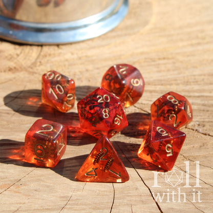 Set of 7 red transparent tabletop roleplaying polyheral dice with gold numbers and copper gears inside on a wooden surface, featuring 'roll with it' logo.