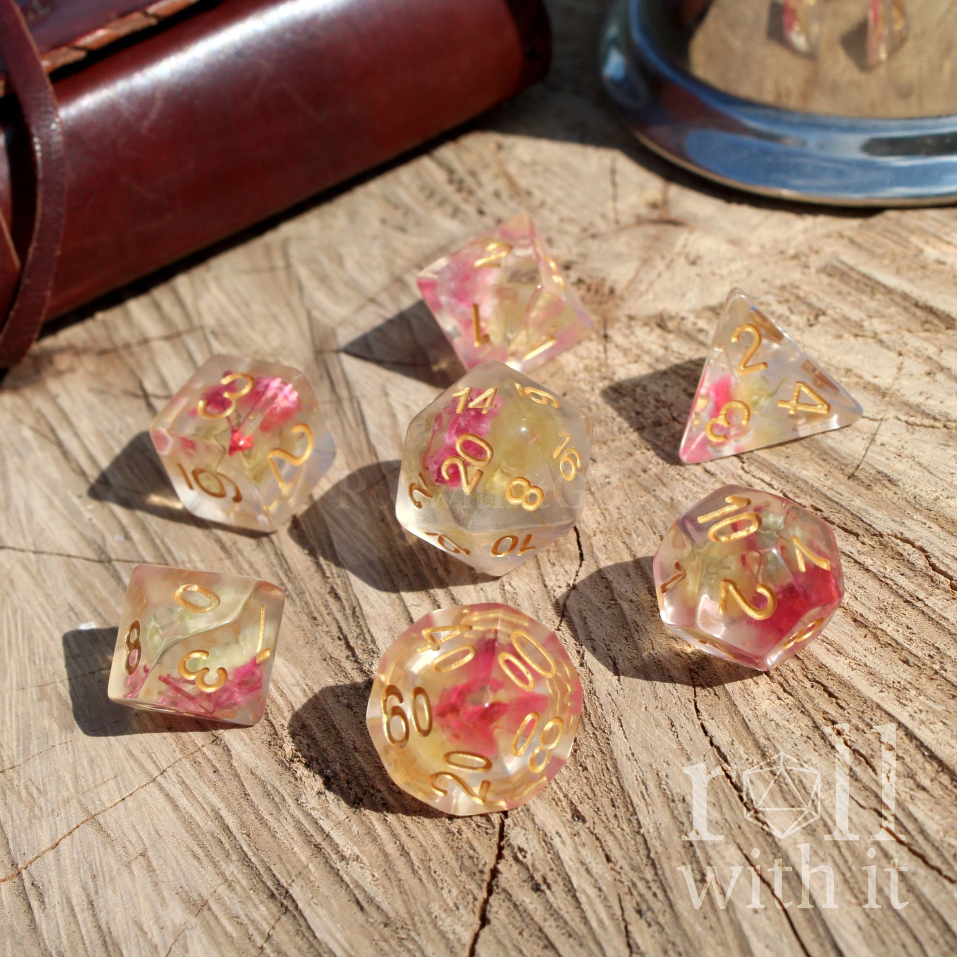 Set of 7 RPG gaming transparent dice with pink and gold flowers and golden numbers on a wooden surface, with 'roll with it' branding.