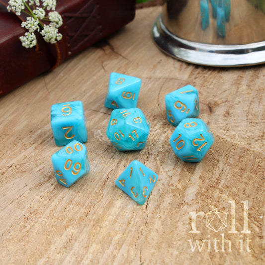 Set of turquoise sailor blue polyhedral dice with gold numbers on a wooden surface, featuring 'roll with it' branding.