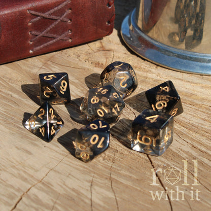 Set of shiny black and gold polyhedral dice on a wooden surface with 'roll with it' logo.