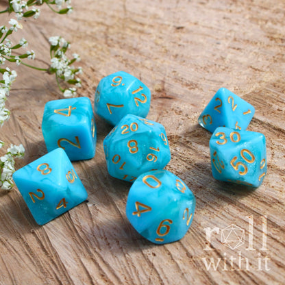 Set of 7 turquoise blue polyhedral dice with gold numbers on a wooden surface, featuring 'roll with it' logo.