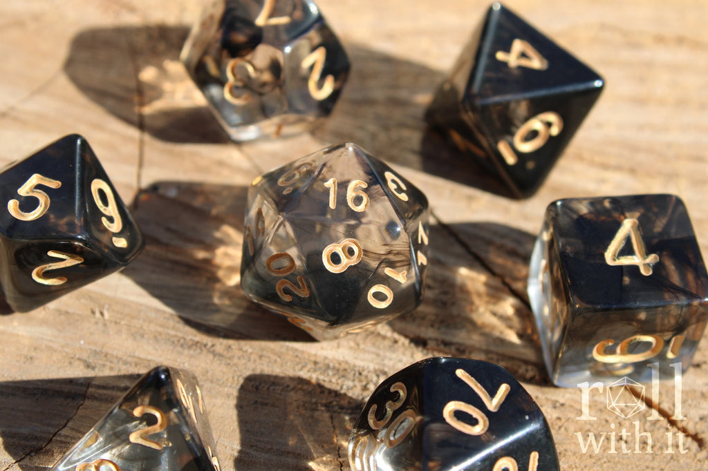 A set of smoky black and clear polyhedral roleplaying dice with gold numbering sitting on an oak surface