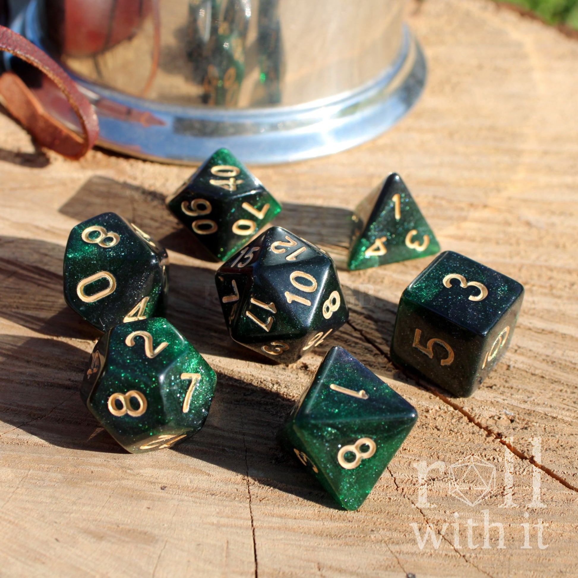 Set of 7 emerald glittery green polyhedral dice with gold numbers on a wooden surface, featuring 'Roll with It' branding.