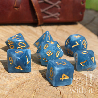 Blue polyhedral dice set with gold numbers on a wooden surface, with a leather book in the background.