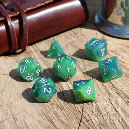 Green blue swirling polyhedral dice set on a wooden surface with a leather-bound book and glass container in the background.
