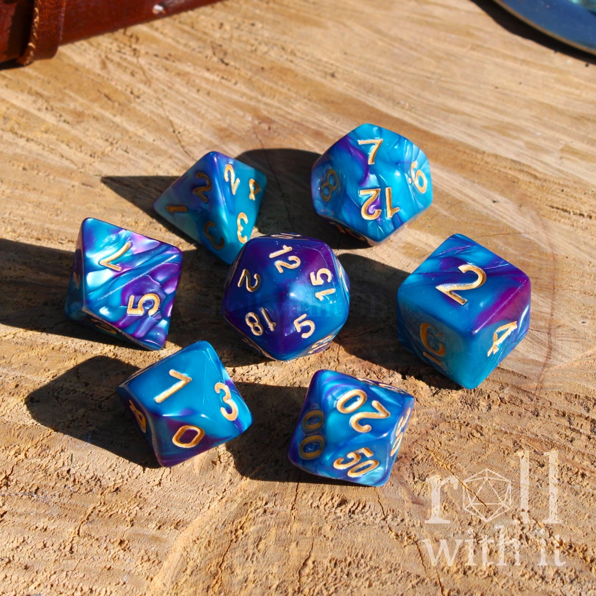 Blue and purple tabletop polyhedral dice set on a wooden surface with 'Roll With It' branding.