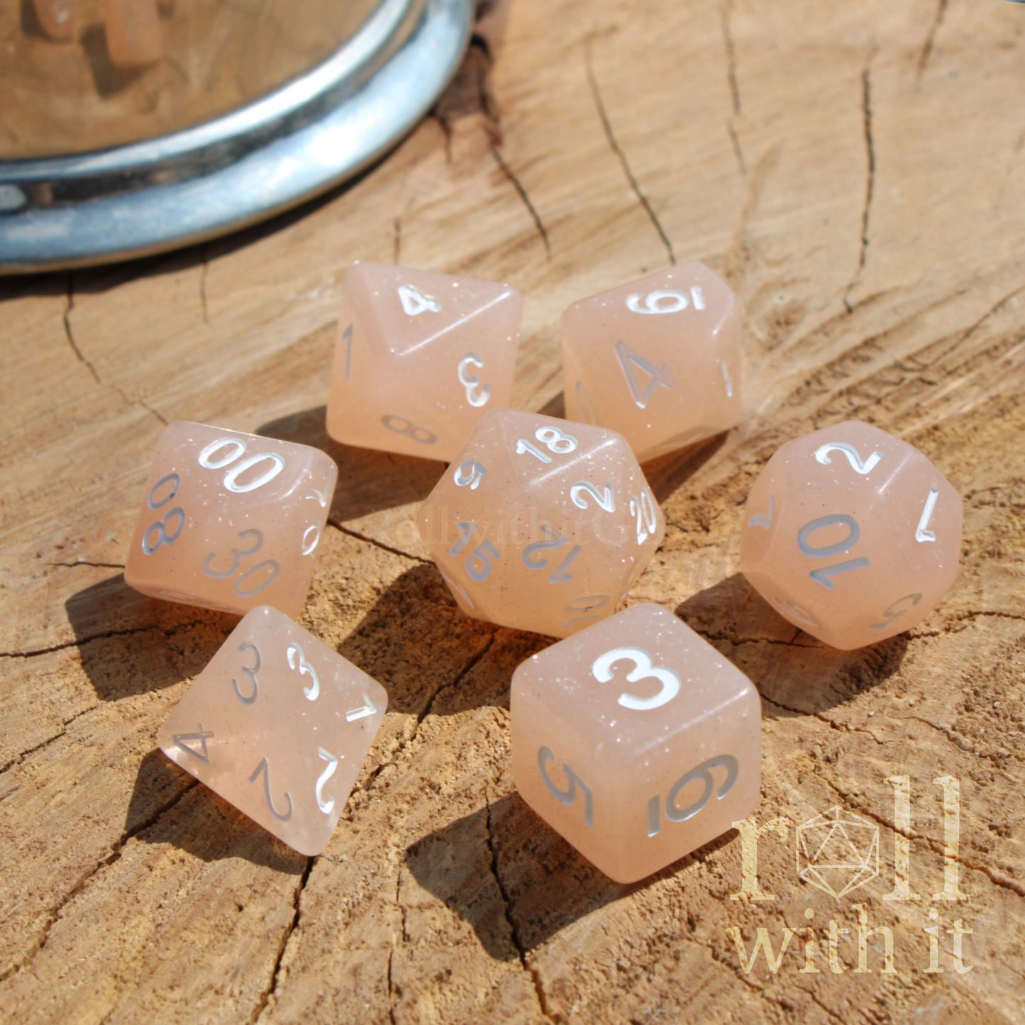 Set of pink beige polyhedral dice with white numbers on a wooden surface, branded 'roll with it'.