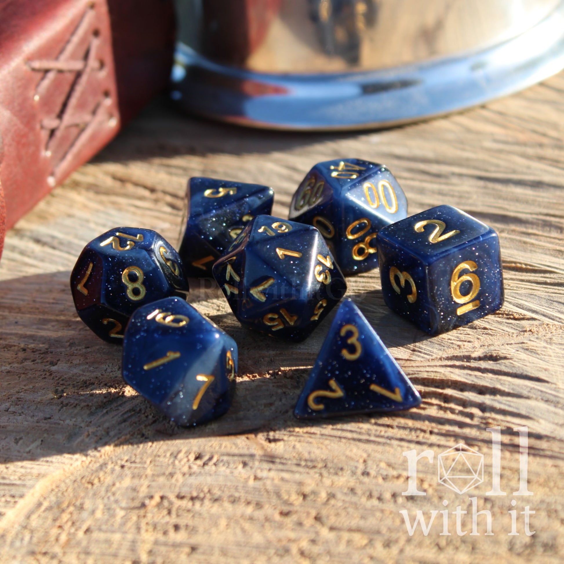 Set of dark mightnight blue starry space polyhedral dice with gold numbers on a wooden surface, featuring the text 'roll with it'.