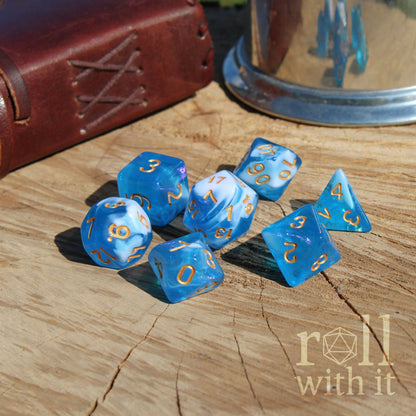Set of blue white sailor themed polyhedral dice with gold numbers on a wooden surface, featuring 'roll with it' logo.