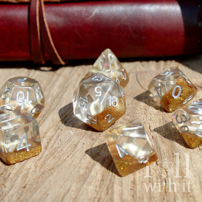 Set of gold and clear sand themed polyhedral dice on a wooden surface with 'roll with it' branding.