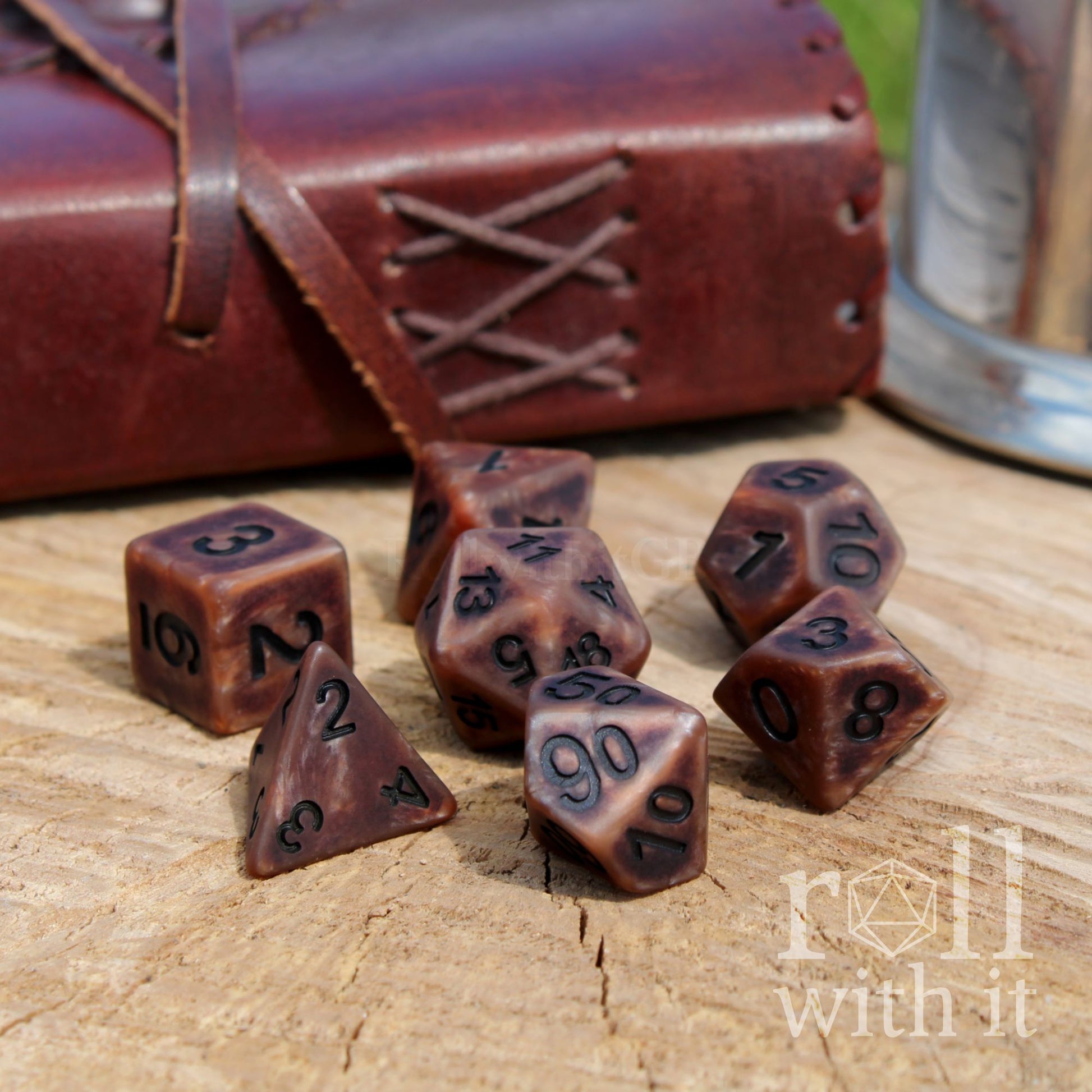 Set of brown bronze polyhedral dice on a wooden surface with a leather-bound book and metal canister in the background.