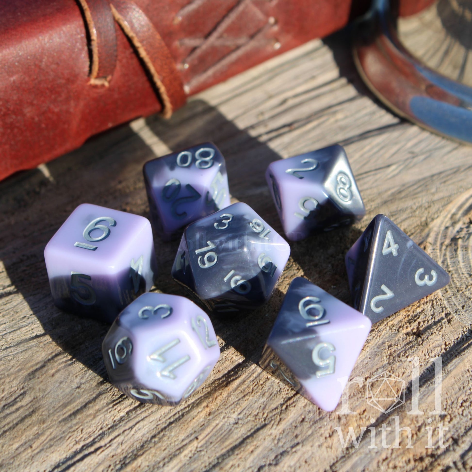 Set of mixed pale lilac purple polyhedral dice on a wooden surface with a leather book in the background.
