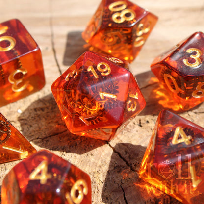 Transparent amber-colored polyhedral dice with gold numbers and copper cogs inside on a wooden surface.