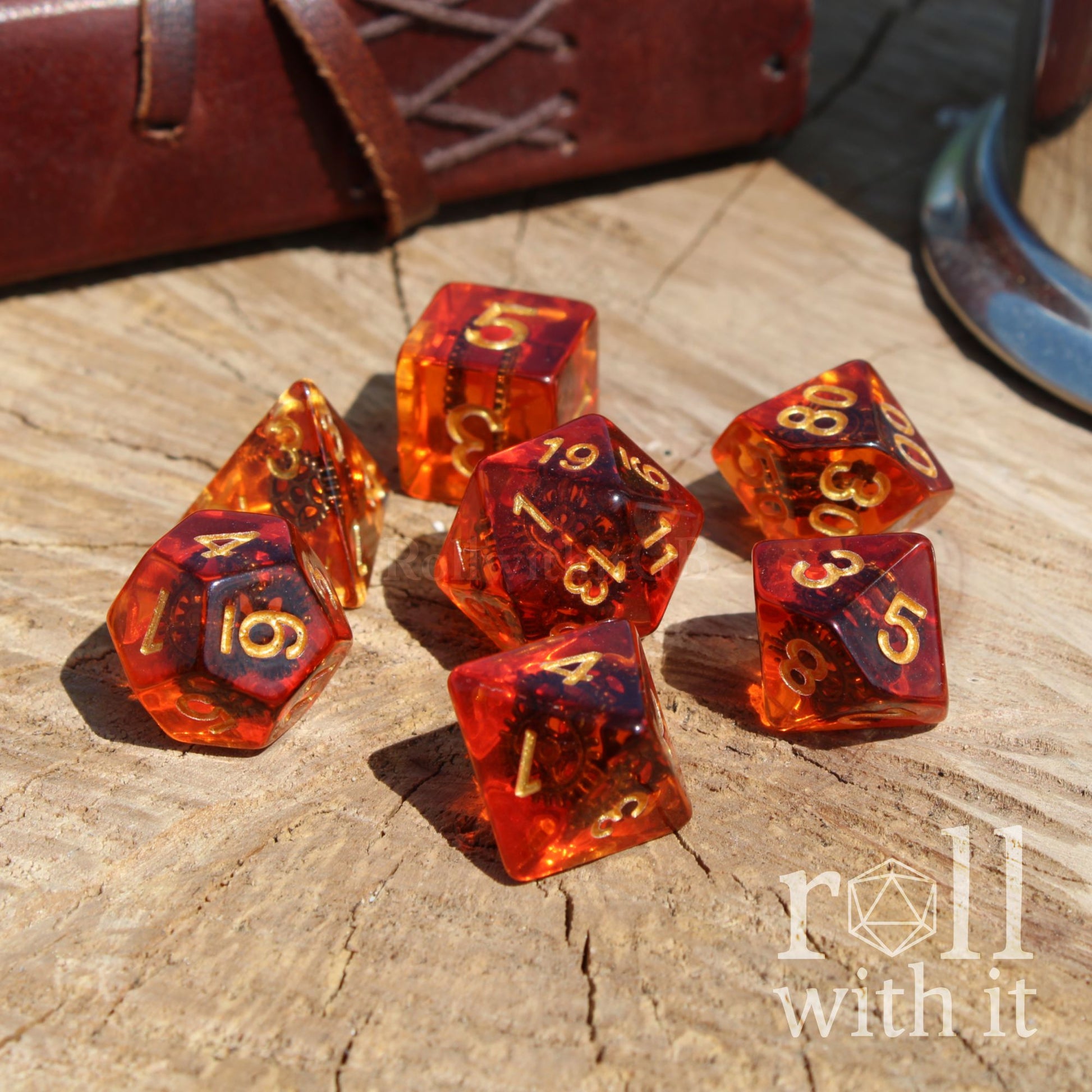 Set of amber-colored polyhedral dice with gold numbers and copper gears on a wooden surface, featuring 'roll with it' branding.
