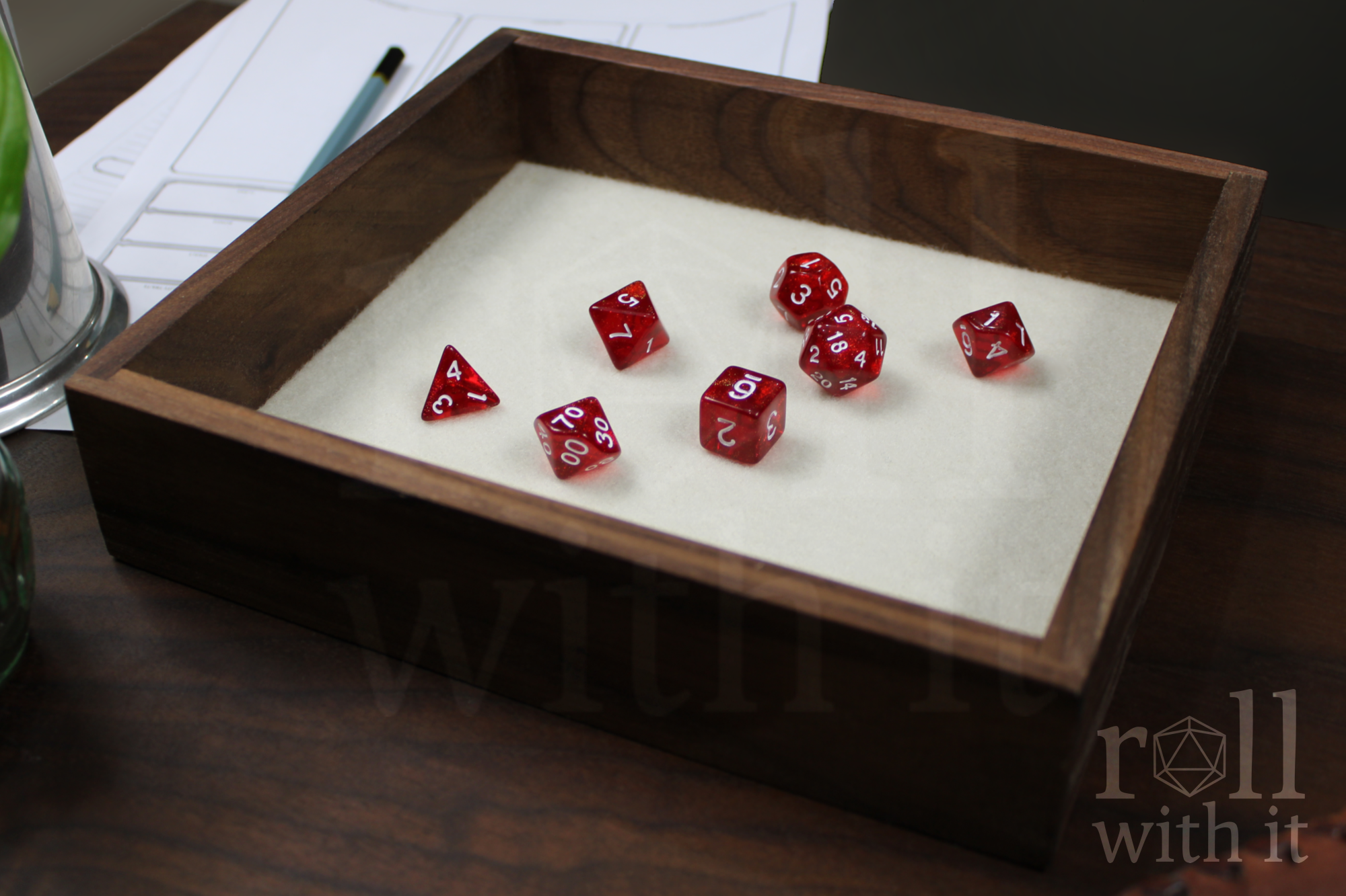 Red dice with white numbers on a wooden walnut dice tray with a cream white felt base, with other tabletop accessories in the background.