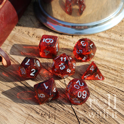 Top view of 7 polyhedral crimson red resin dice swirling and galaxy themed on a wooden surface