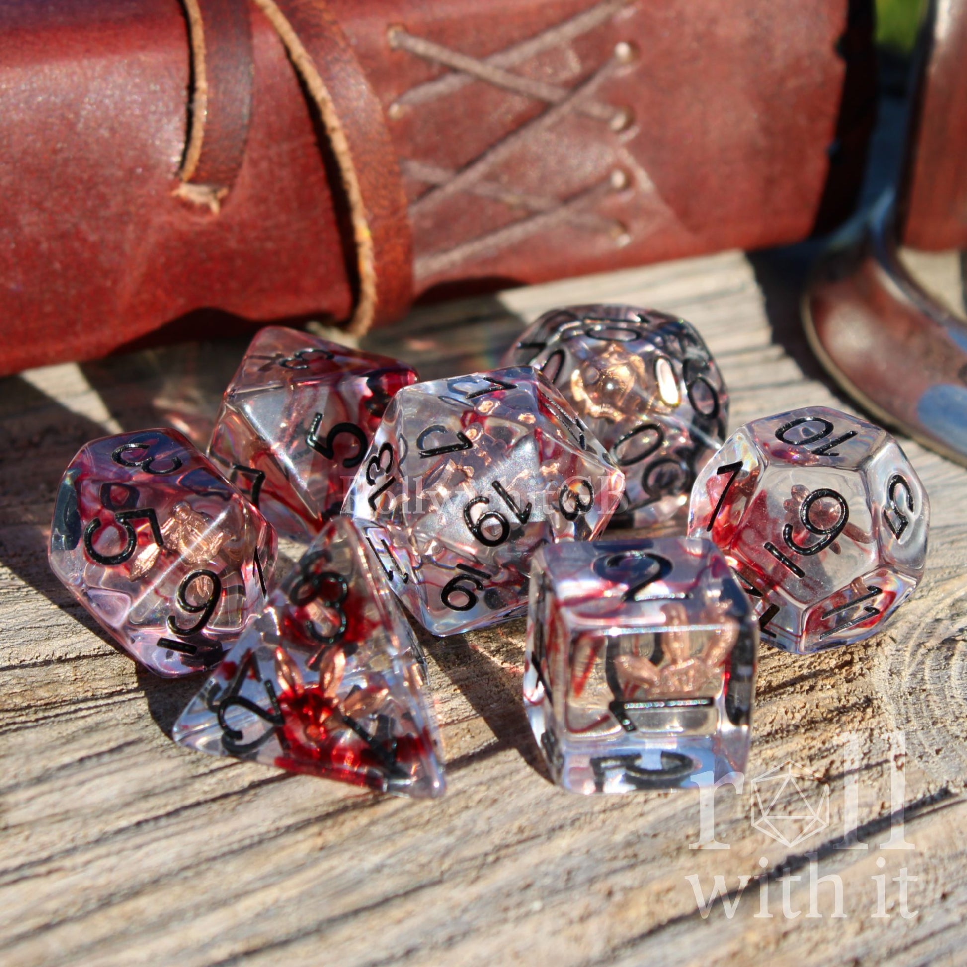 Set of transparent dice with blood red swirls and black numbers and rose gold 3 daggers inside on a wooden surface.