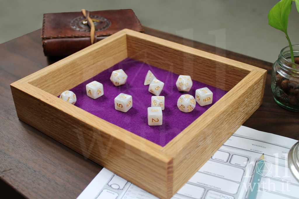 Wooden red oak dice tray with tyrian purple felt interior on a walnut table. A transparent 'roll with it' logo sits in the right side. White pearl dice sit inside the box on the felt base. Other tabletop accessories are in the background.