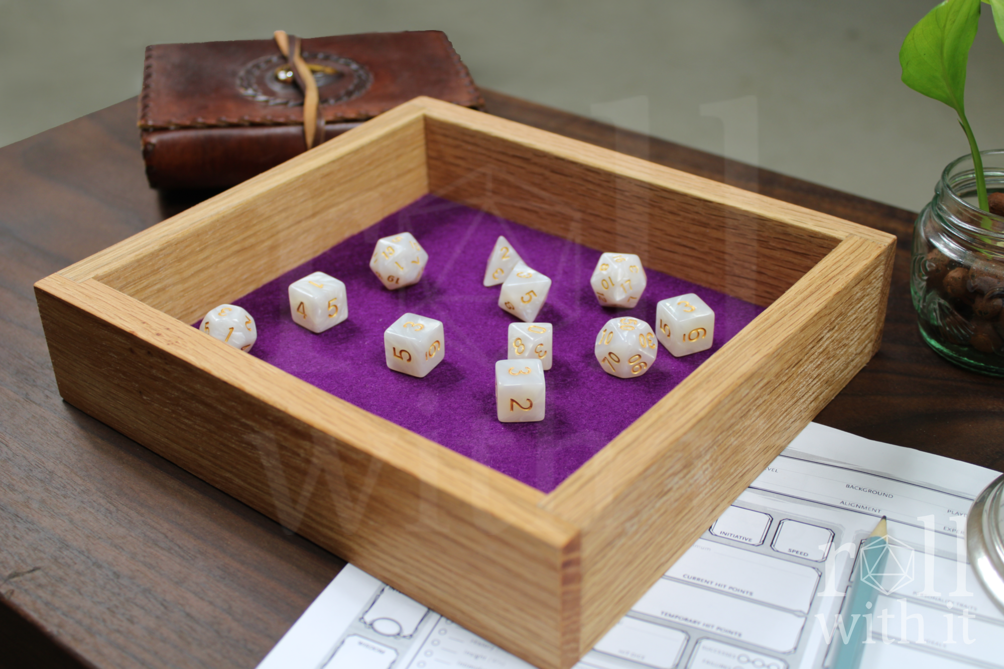 Wooden red oak dice tray with tyrian purple felt interior on a walnut table. A transparent 'roll with it' logo sits in the right side. White pearl dice sit inside the box on the felt base. Other tabletop accessories are in the background.