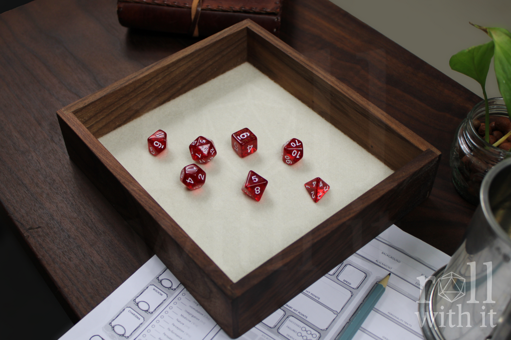 Solid walnut roleplaying dice tray with red DnD polyhedral dice, sat on a white felt base. A character sheet and other tabletop accessories are in the background.