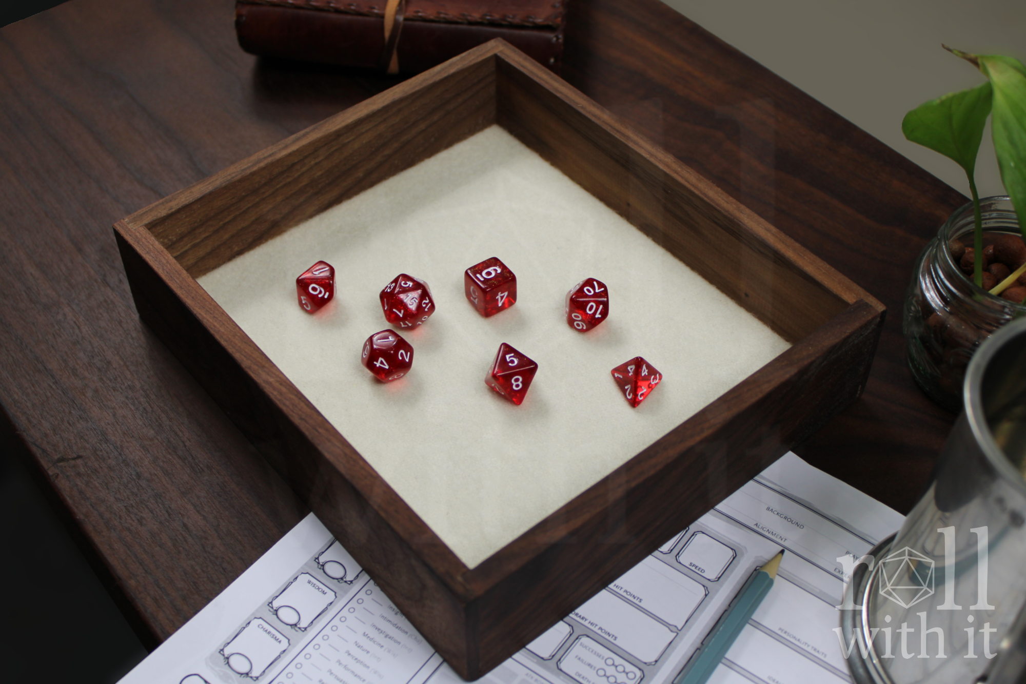 Solid walnut roleplaying dice tray with red DnD polyhedral dice, sat on a white felt base. A character sheet and other tabletop accessories are in the background.