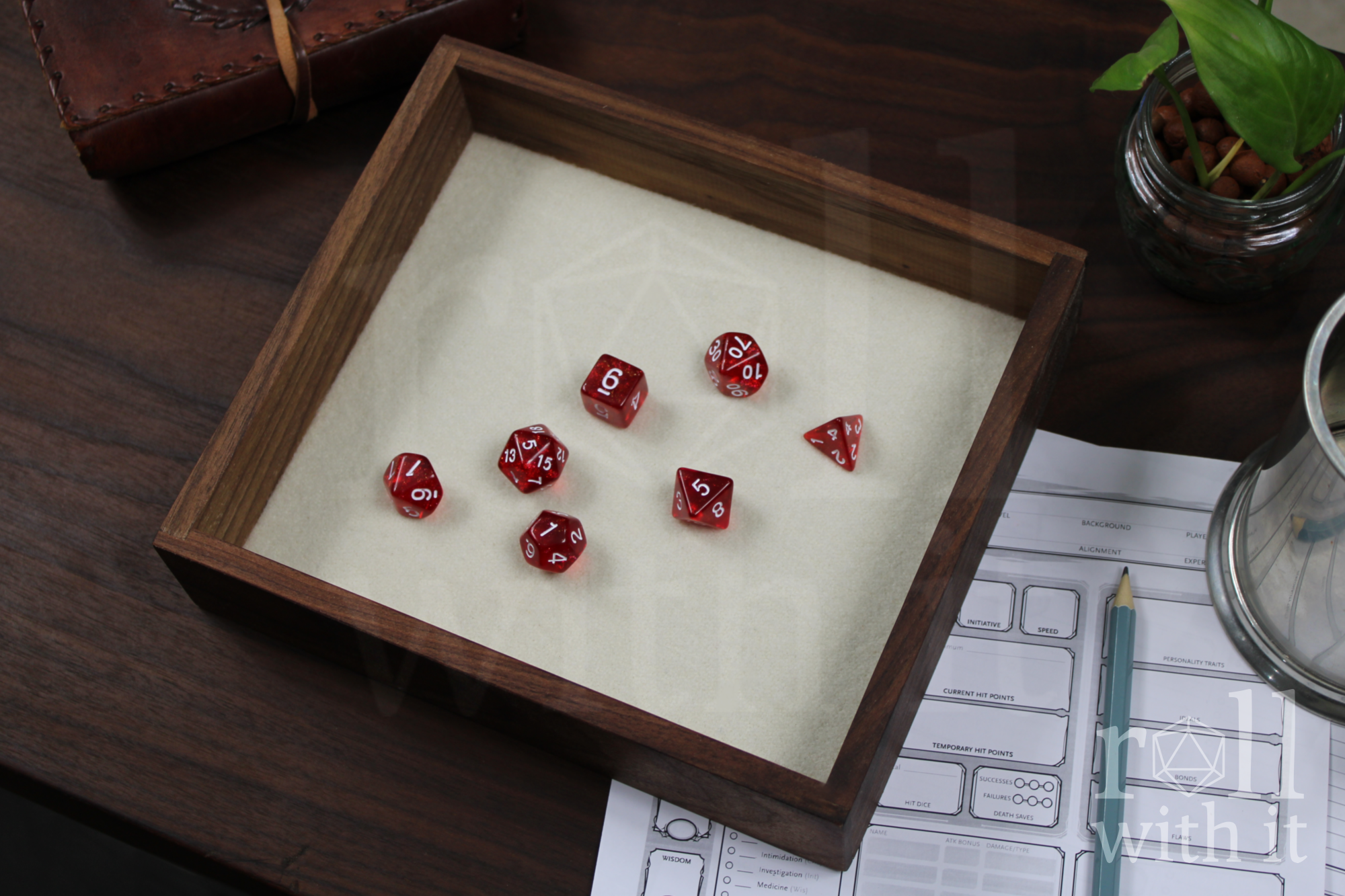 Red dice with white numbers on a wooden walnut dice tray with an ivory white felt base, with other tabletop RPG accessories in the background.