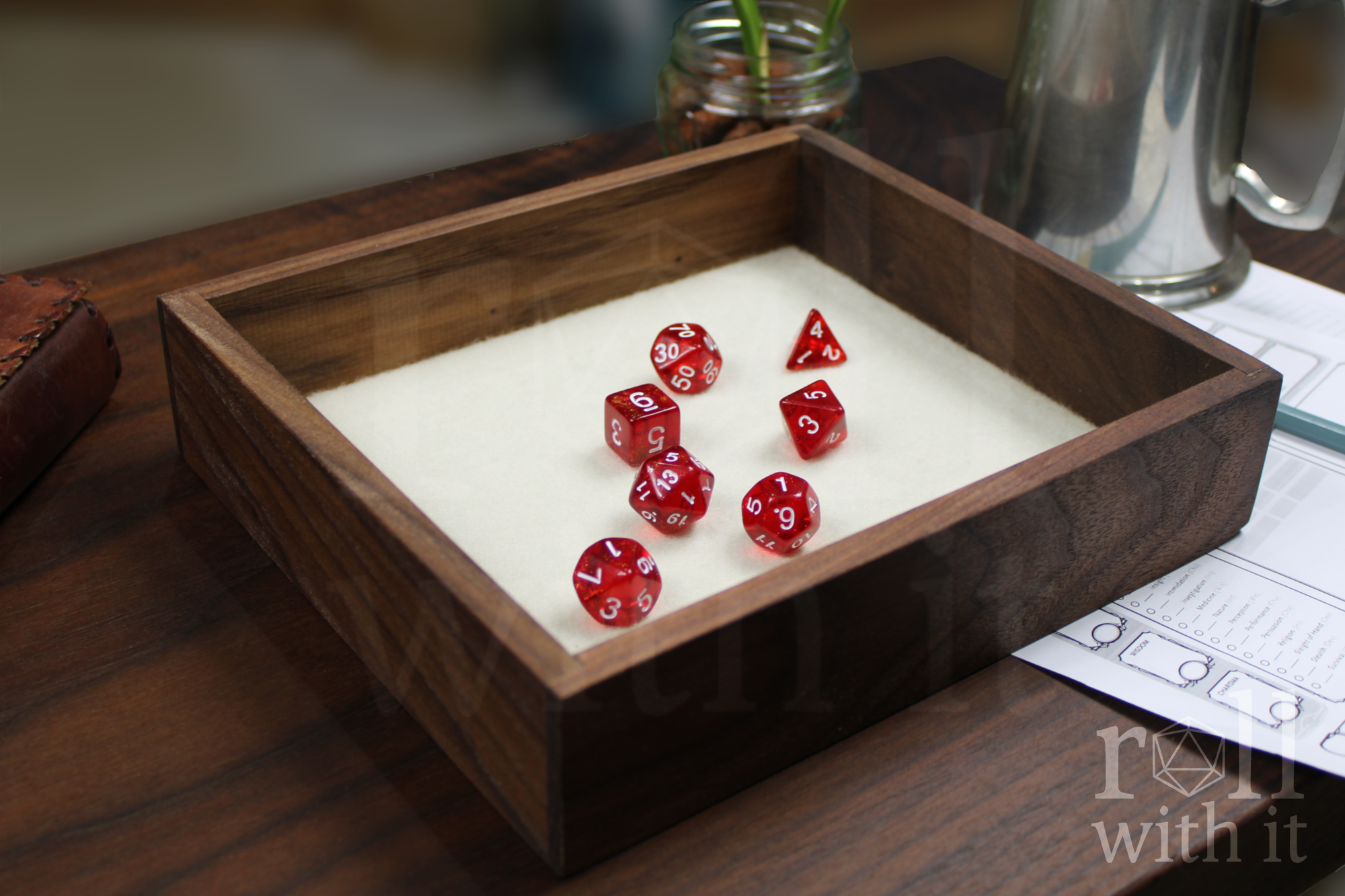 Red dice with white numbers on a wooden walnut dice throwing tray, on a white felt base, with other tabletop accessories in the background.