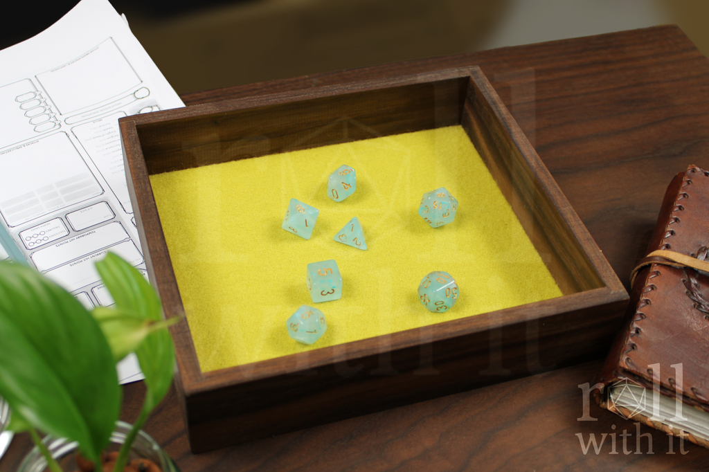 Wooden dice tray with blue dice on a yellow cloth, next to a plant and notebook.