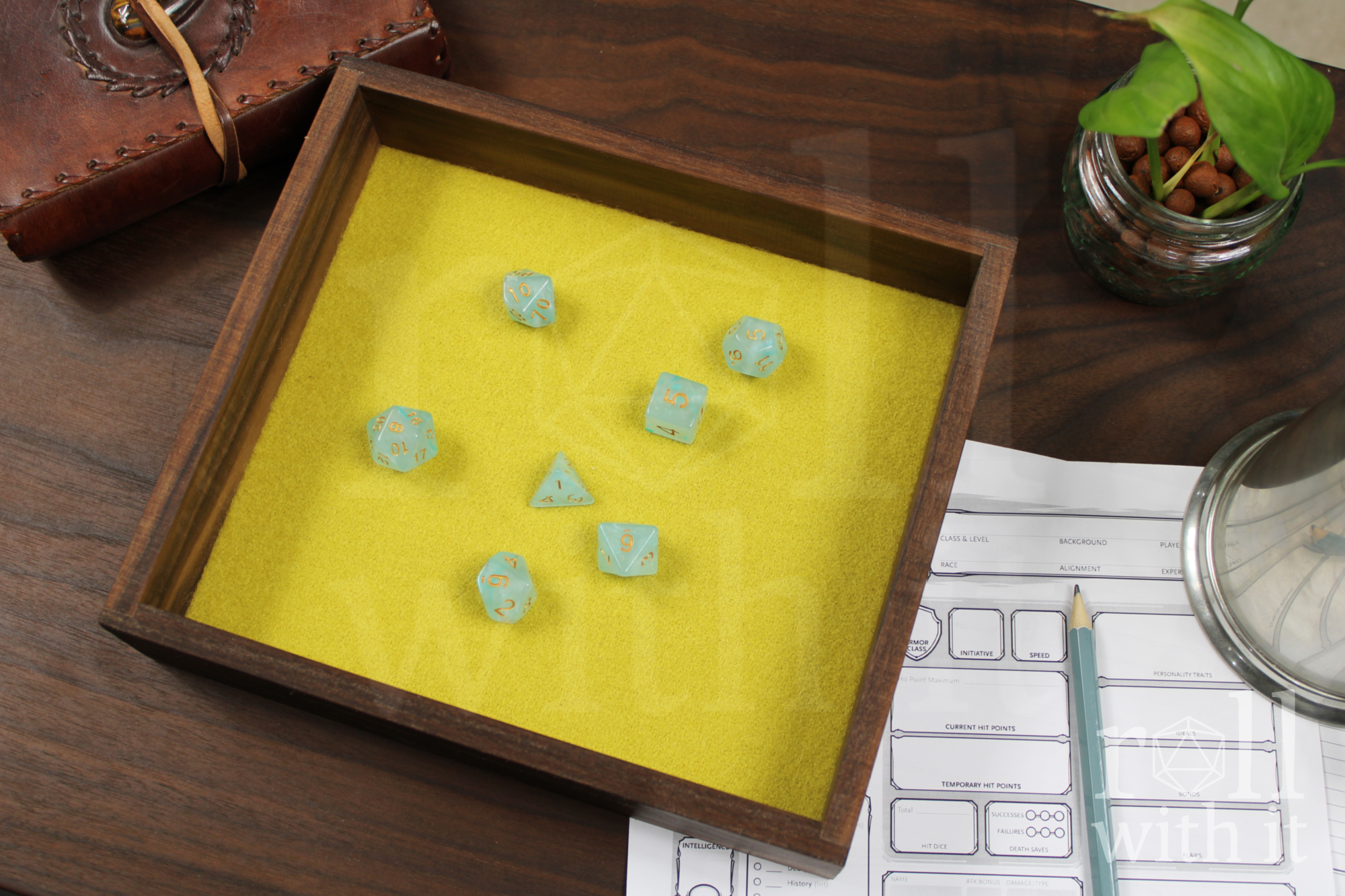 Wooden walnut dice tray with clear blue dice on a yellow felt base, next to a leather notebook and other dungeons and dragons accessories.