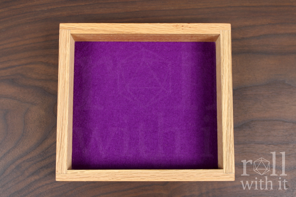 Wooden red oak dice tray with tyrian purple wool felt base interior on a wooden walnut table. A 'roll with it' logo sits in the bottom right corner.