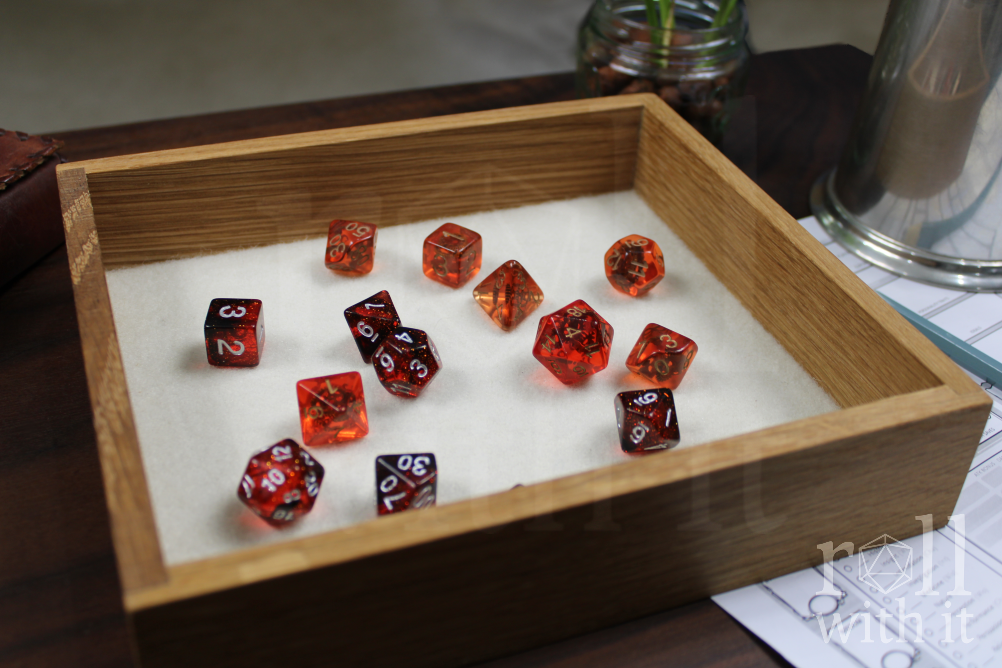 Orange, Red and black dungeons and dragons tabletop RPG dice set sat on a white cream felt base in a wooden white oak dice tray on a table. The 'roll with it' logo is in the corner.