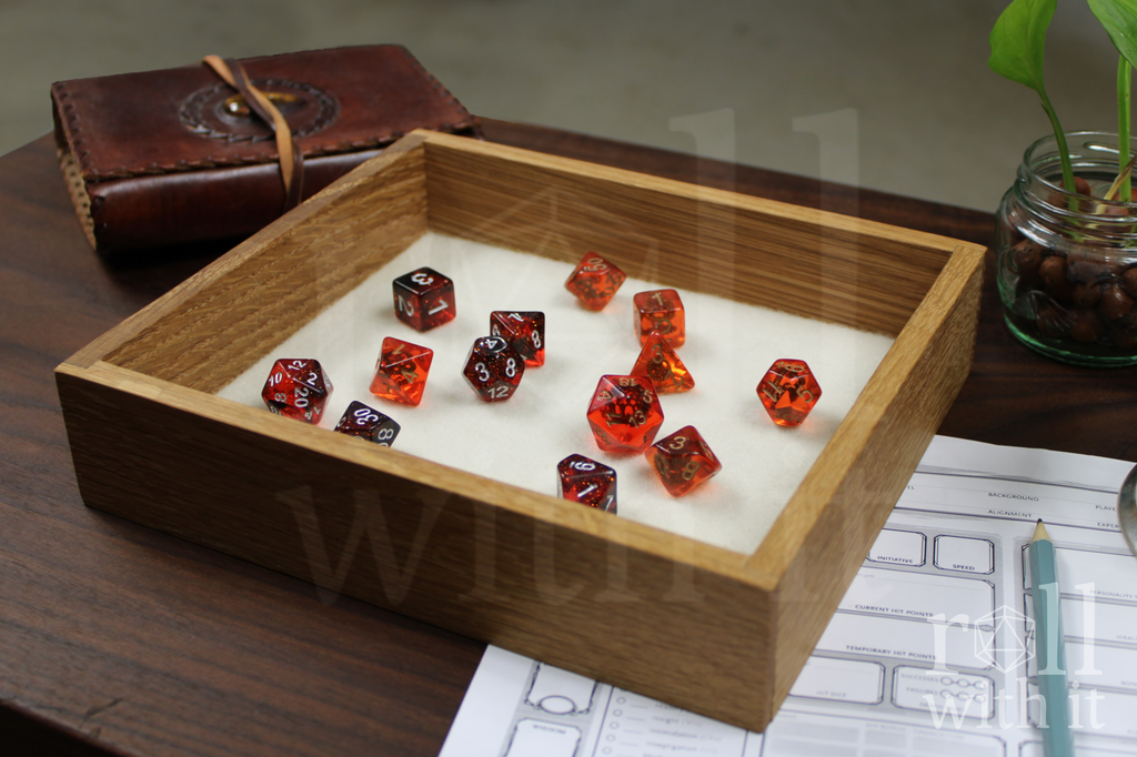 Red and black DnD tabletop roleplaying dice sat on a white cream felt base in a solid wooden white oak dice tray with 'roll with it' branding in the corner of the image. A character sheet and leather journal are in the backround.