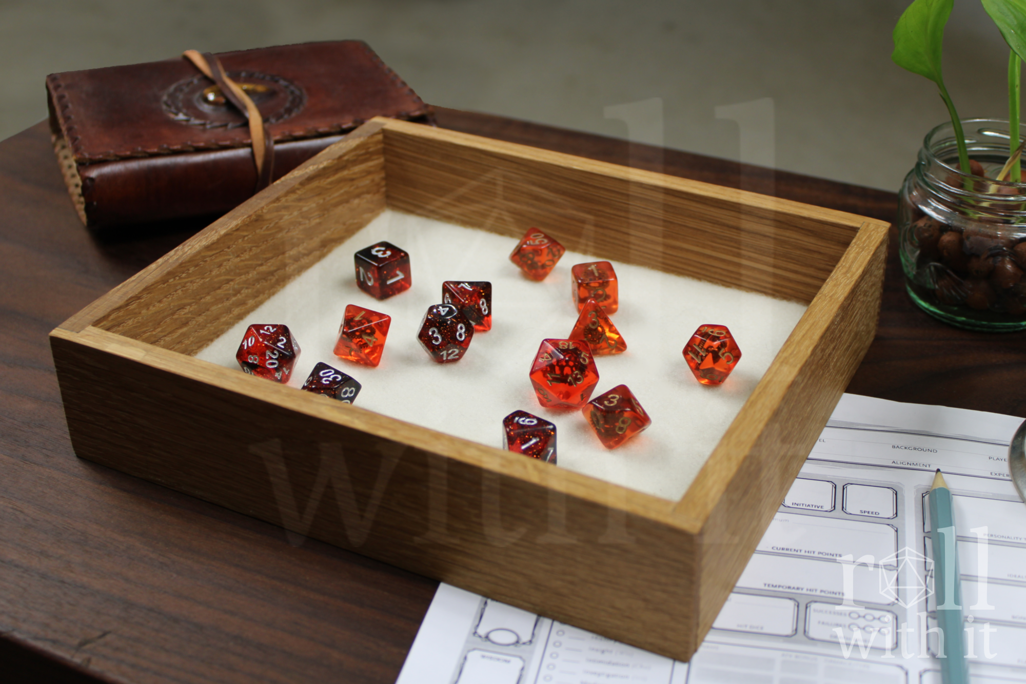Red and black DnD tabletop roleplaying dice sat on a white cream felt base in a solid wooden white oak dice tray with 'roll with it' branding in the corner of the image. A character sheet and leather journal are in the backround.