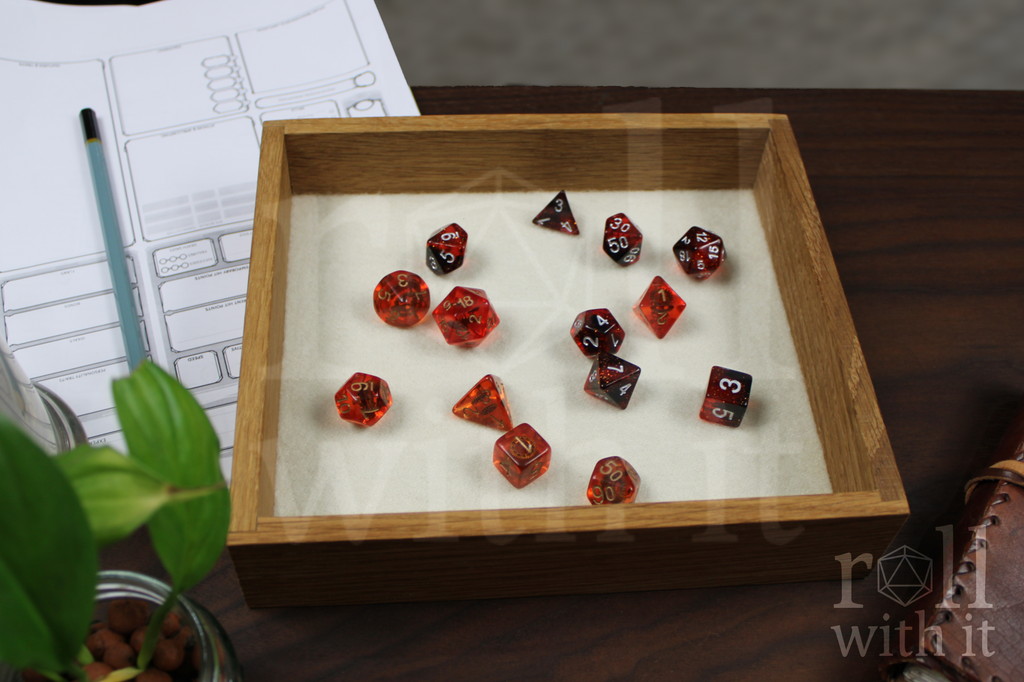 Orange, Red and black Dungeons and Dragons RPG dice sat on a cream white felt base in a wooden white oak tabletop dice tray on a table with 'roll with it' branding.