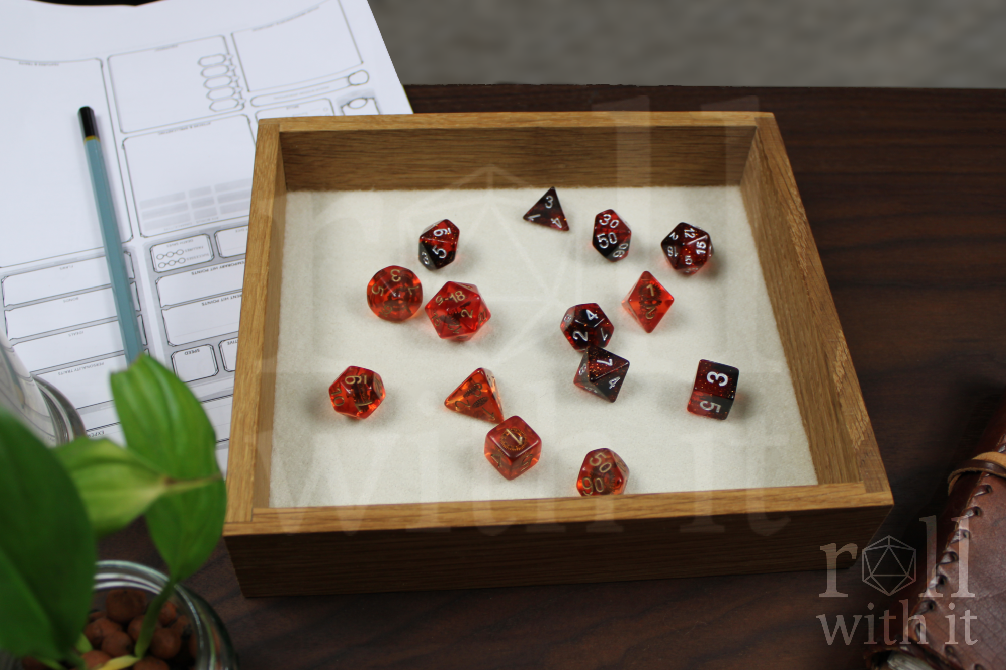 Orange, Red and black Dungeons and Dragons RPG dice sat on a cream white felt base in a wooden white oak tabletop dice tray on a table with 'roll with it' branding.