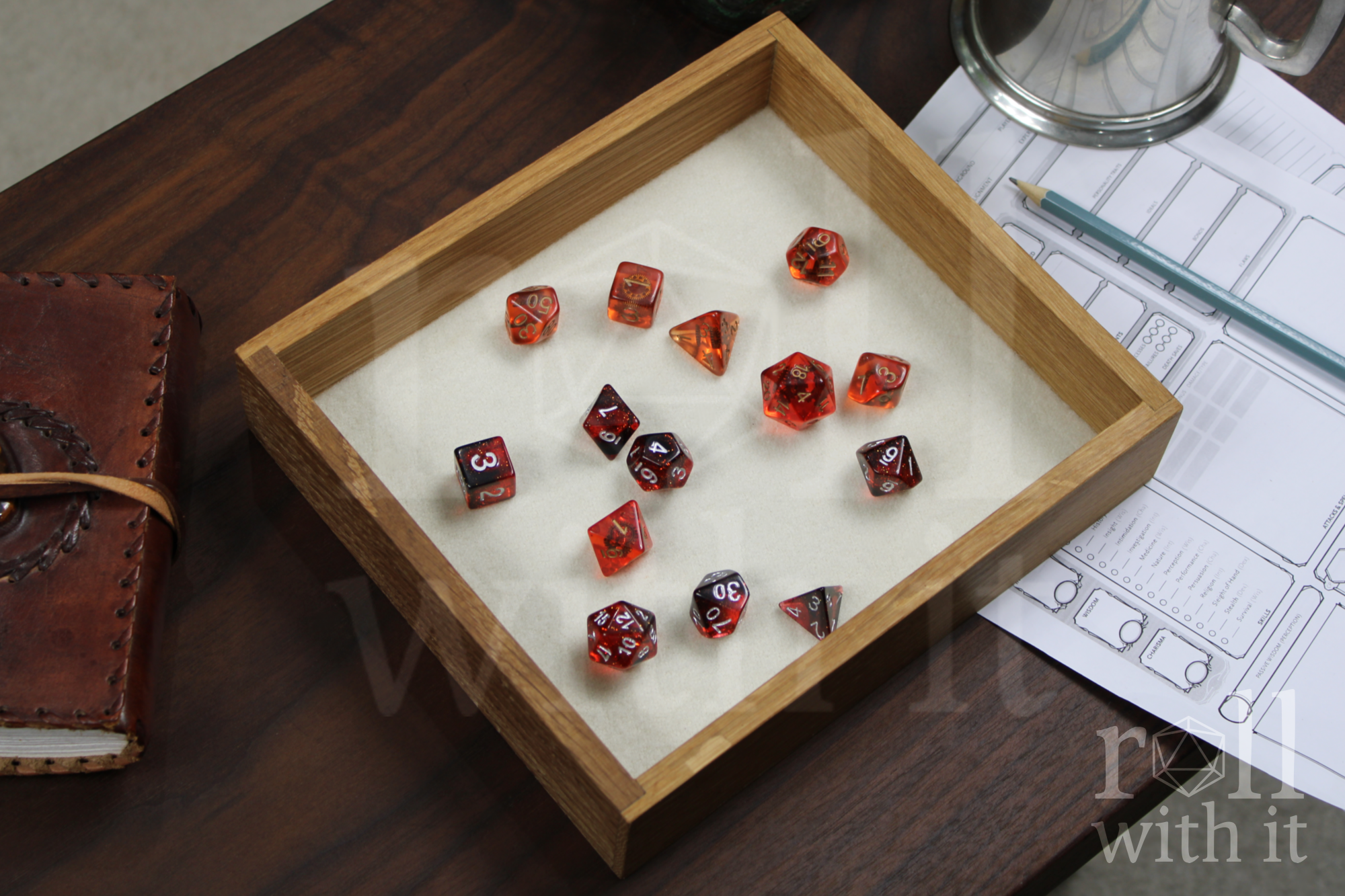 Orange, Red and black Dungeons and Dragons RPG dice sat on a cream white felt base in a wooden white oak tabletop dice tray on a table, surrounded by character sheets and a leather bound journal.